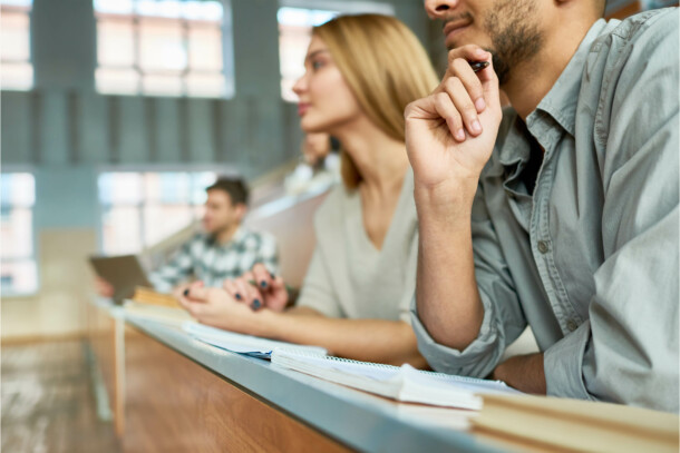 Multi-ethnic group of students sitting at desk in lecture hall of modern college and listening to professor, focus on young Middle-Eastern man, copy space