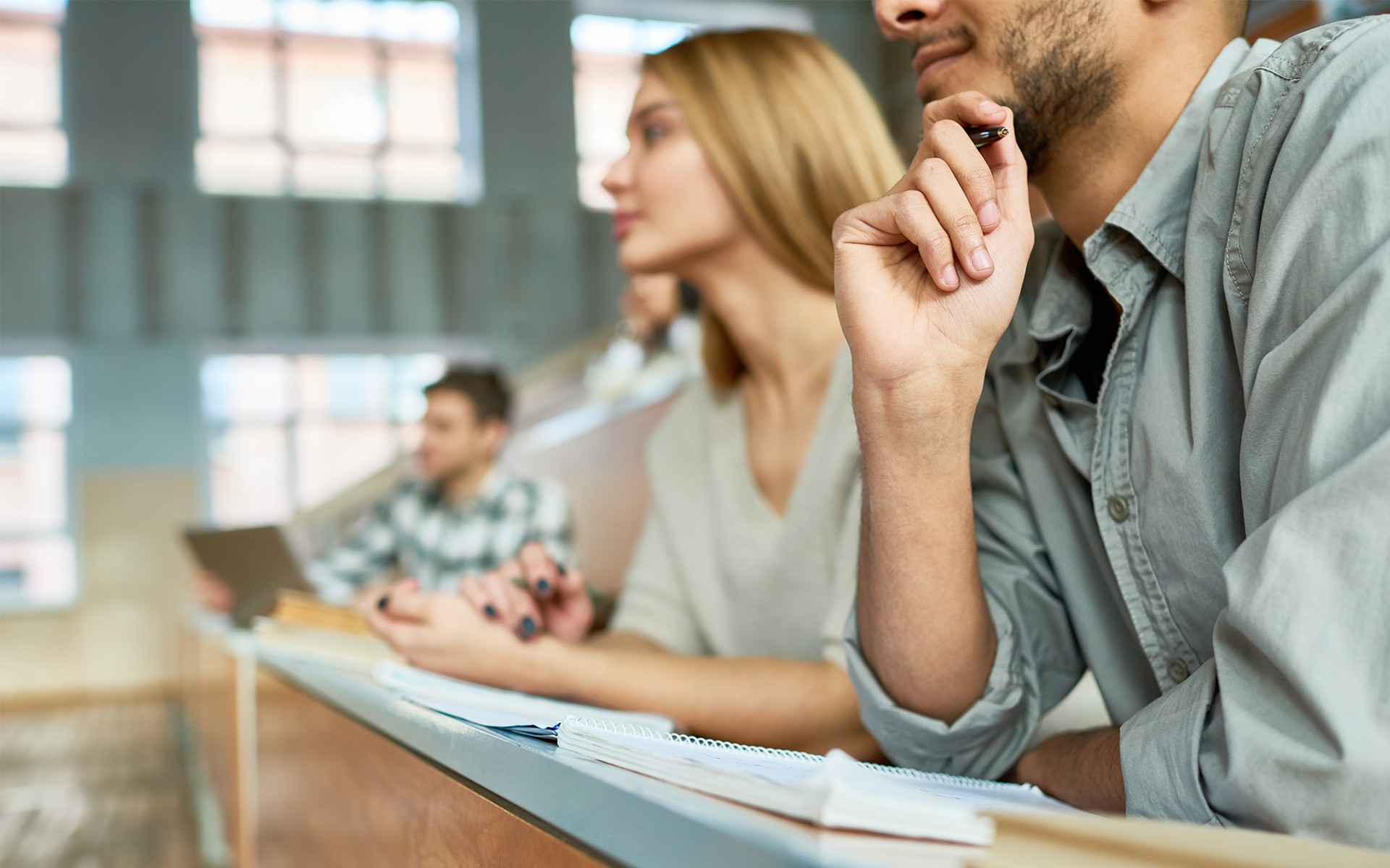 Multi-ethnic group of students sitting at desk in lecture hall of modern college and listening to professor, focus on young Middle-Eastern man, copy space